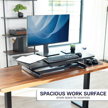 Wooden desk with an adjustable standing desk converter on it, computer monitor, keyboard, and mouse in a home office setting.