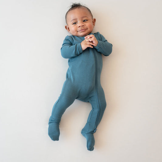 Infant smiling with hands clasped together wearing the Ribbed Zipper Footie in Atlantic on a bisque blanket
