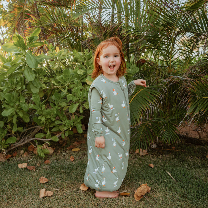 Young girl standing in a field touching a plant wearing the Sleep Bag Walker in Silly Goose 2.5 with long sleeve toddler pajamas on underneath