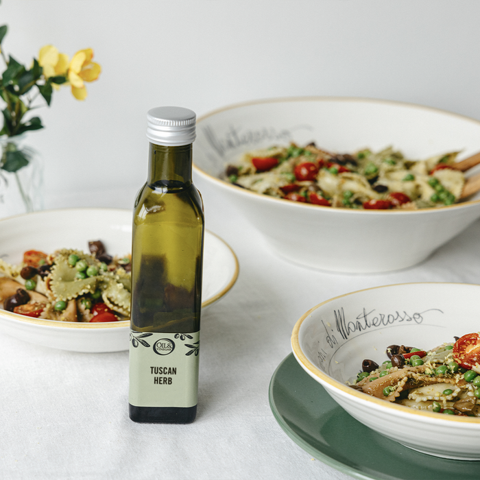 Tuscan Herb flavored olive oil bottle on a white table next to bowls of pasta salad